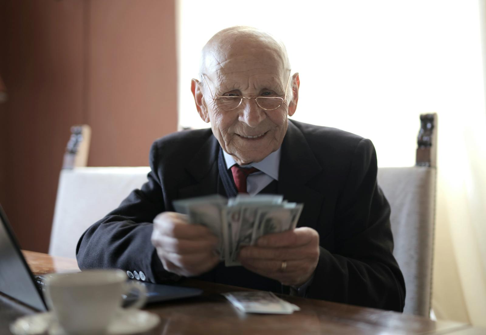 Positive senior businessman in formal suit and eyeglasses counting money bills while sitting at wooden table with cup of beverage and near opened laptop